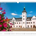 Im Juni-Kalenderbild: Rathaus mit Glockenspiel, blauem Himmel und blühenden pinken Blumen im Vordergrund.