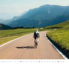 Text: "Juni". Ein Radfahrer auf einer kurvigen Straße in einer bergigen Landschaft bei sonnigem Wetter. Logo unten rechts.