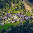 Eine idyllische Berglandschaft mit einem Dorf in der Mitte, umgeben von grünen Wiesen und dichten Wäldern.