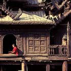 Kein Text erkennbar. Ein Mönch in roter Robe sitzt in traditioneller Holzarchitektur mit geschnitzten Details.