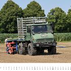 JUNI. Ein Unimog zieht landwirtschaftliche Geräte über ein Feld. Im Hintergrund sind grüne Bäume zu sehen.
