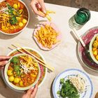 Text: "Laksa-Party (Seite 90)". Verschiedene Schalen mit buntem Laksa, Garnelen, Eiern, Kräutern und Knusperstreifen. Hände servieren.