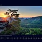07 Juli. Kalender mit Landschaft: Sonnenuntergang über einem grünen Tal, Felsen und Kiefern im Vordergrund.