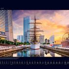 06 Juni. Ein Segelschiff spiegelt sich im Wasser, umgeben von modernen Gebäuden und einem großen Riesenrad bei Sonnenuntergang.