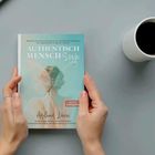 "AUTHENTISCH MENSCH Sein" auf einem Buch gehalten von zwei Händen, daneben eine Tasse Kaffee.