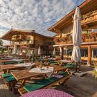 Rustikale Holzgebäude mit Blumenkästen, Holztische mit weißen Tischdecken, grüne Polster, Sonnenschirme und blauer Himmel.