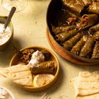 Weinblätter gefüllt mit Reis, in einer Pfanne. Zitronenscheiben, Brot und Joghurt serviert. Ein Glas Weißwein daneben.