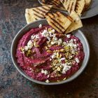 Ein Teller mit violettem Dip, garniert mit zerbröckeltem Feta und Pistazien, daneben gegrillte Pita-Dreiecke.