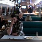 Keine sichtbaren Texte. Ein Mann sitzt nachdenklich in einem Zugrestaurant. Weitere Passagiere im Hintergrund.