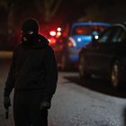 Ein Mensch in schwarzer Kleidung und Skimaske steht nachts auf einer Straße. Autos mit eingeschaltetem Rücklicht im Hintergrund.
