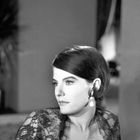 Eine Frau mit kurzen Haaren und Ohrringen, trägt ein Spitzenoberteil und schaut zur Seite. Sie wirkt elegant und konzentriert.