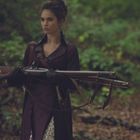 Keine sichtbaren Texte. Eine Person im lila Mantel hält zwei Gewehre in einem Wald.