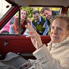 Eine ältere Frau sitzt in einem roten Auto, während drei Personen durch das Fenster neugierig hereinblicken.