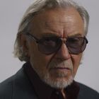 Ein älterer Mann mit grauen Haaren und Bart trägt eine Sonnenbrille und einen dunklen Anzug.