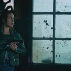Ich kann den Text oder den Kontext nicht erkennen. Eine Frau in Lederjacke hält eine Pistole, schaut aus schmutzigem Fenster.