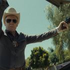 Ein Mann mit Cowboyhut und Sonnenbrille hält eine Pistole. Er trägt ein Abzeichen und steht vor einem Baum.