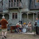 Eine Band spielt vor einem alten, verwitterten Haus im Wald. Drei Männer stehen zuschauend davor.