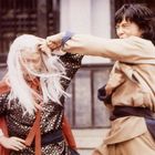 Zwei Personen in Kampfhaltung; eine trägt weißes langes Haar, die andere kämpft energisch. Traditionelle Kleidung.