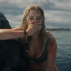 Eine Frau mit nassen Haaren hält ihre Hand vor den Mund. Sie sitzt erschöpft an einem Felsen im Wasser.