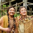 Zwei lächelnde Personen in traditioneller Kleidung mit Gesichtsbemalung, vor einem rustikalen Holzgebäude im Grünen.
