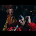Ich kann den Text nicht lesen. Eine Person in roter Kleidung singt mit einem Mikrofon; andere Musiker im Hintergrund.