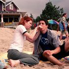 Ein Mann und eine Frau sitzen am Strand auf einer Decke vor einem Haus. Sie scheinen miteinander zu interagieren.