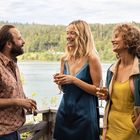 Drei Personen stehen lachend auf einer Terrasse mit Weingläsern, vor einem See und Wald im Hintergrund.