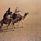 Drei Personen reiten auf Kamelen durch eine trockene, rissige Wüstenlandschaft.