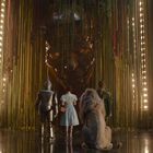 Vier Figuren stehen vor einem riesigen, beleuchteten, imposanten Gesicht in einem von Lichtern umrahmten Raum.