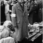 Ein Mann mit Stern-Armbinde steht vor Koffern. Neben ihm eine Menschengruppe und Soldaten in Uniform.