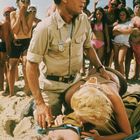 Ein Mann mit Abzeichen kniet im Sand neben einer Frau, die einen leblosen Körper beatmet, umgeben von einer Menschenmenge am Strand.