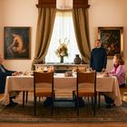 Ein Tisch in einem eleganten Raum, gedeckt zum Essen. Drei Personen, zwei sitzend, eine steht. Wände mit Gemälden, Teppich am Boden.