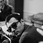 Ein Mann mit Sonnenbrille schaut zur Seite, während ein anderer Mann im Hintergrund sitzt. Alles in einem Bürosetting.