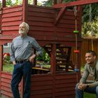 Zwei Männer entspannen sich neben einem roten Holzspielhaus im Garten, einer steht, der andere sitzt auf einer Schaukel.