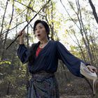 Keine sichtbaren Texte. Eine Frau im blauen Kleid steht im Wald, hält einen Stock und ein Buch, sieht überrascht aus.