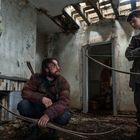 Ein heruntergekommenes Zimmer mit offenem Dach. Zwei Personen mit Bögen, eine kniend, die andere stehend, sind in verwahrloster Kleidung.