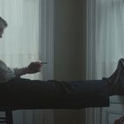 Zwei Männer sitzen entspannt in einem Raum, einer mit Zigarette, die Beine lässig ausgestreckt. Weiches Licht kommt durch Vorhänge.