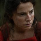 Keine erkennbaren Texte. Eine Frau mit ernstem Gesichtsausdruck, unordentliches Haar, trägt ein rotes Oberteil.