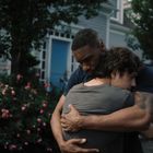 Zwei Menschen umarmen sich vor einem Haus mit blauer Tür, umgeben von rosa blühenden Sträuchern.