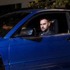 Ein Mann mit ernster Miene sitzt in einem blauen Auto auf dem Fahrersitz, umgeben von dunkler Umgebung und Bäumen.
