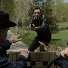 Ein Mann mit Waffe zielt auf drei Personen auf der Ladefläche eines Fahrzeugs, umgeben von Bäumen.