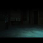 Zwei Menschen stehen in einem dunklen Raum, der von Bodennebel bedeckt ist. Eine Person blickt in einen Spiegel.