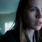 Eine Frau mit langen blonden Haaren schaut ernst in die Ferne. Im Hintergrund ist eine unscharfe Gestalt zu sehen.