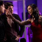 Eine Frau in einem roten Kleid zeigt auf zwei Männer in schwarzen Anzügen vor lila beleuchtetem Hintergrund.