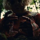 Person in dunklem, unheimlich wirkendem Outfit mit blutigen Händen im Wald; heller Hintergrund mit Bäumen.