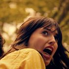 Keine Texte sichtbar. Eine Frau in Gelb schaut erschrocken auf etwas. Im Hintergrund Bäume in warmen Farben.