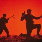Zwei Soldaten in Silhouette. Einer steht mit Gewehr, der andere kniet mit Fernglas. Hintergrund leuchtend rot.