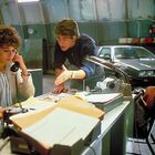 Eine Frau am Telefon; ein Mann beugt sich über den Tisch, gestikuliert. Im Hintergrund ein graues Auto in einer Halle.