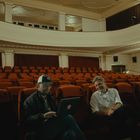 Zwei Personen sitzen entspannt in einem leeren Kinosaal. Der eine nutzt einen Laptop, der andere schaut lächelnd zu.