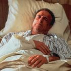 Ein Mann liegt in einem Bett mit einem Gipsarm. Er trägt gestreifte Pyjamas, umgeben von weißen Kissen und einer Blumendecke.